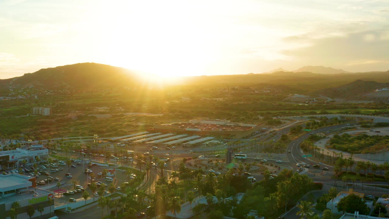 Opening aerial frame over San Jose del Cabo roads and parking areas with the low sun flaring above dark hills and no visible people.