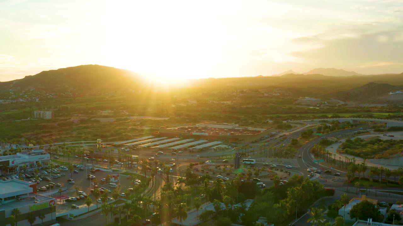 Sunset aerial of highway lanes, traffic, parking lots, trees, and distant mountains.