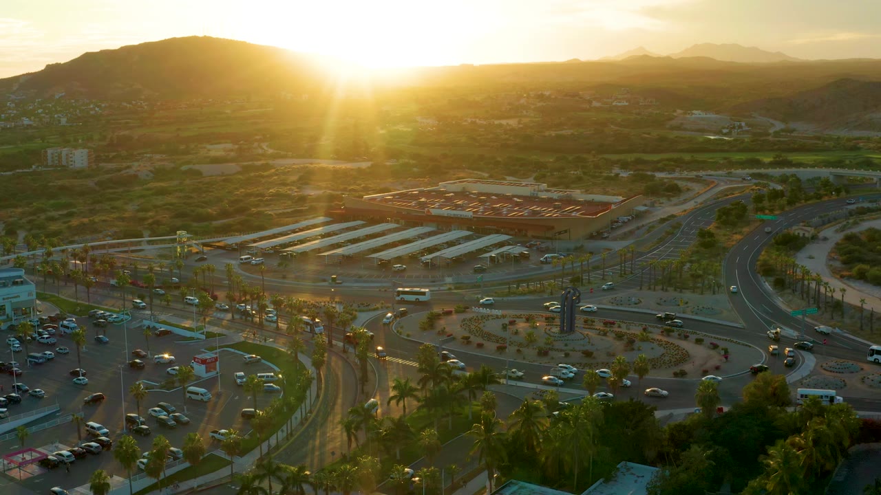 San Jose del Cabo roundabout traffic continues below the drone view, with a bright sunbeam over the hills and no visible people in the roadway scene.