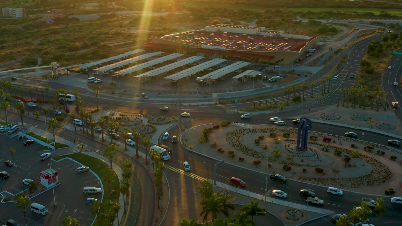 Aerial view over a landscaped roundabout in San Jose Del Cabo, Baja California Sur, with cars circulating, a parking area beyond the road, warm sun glare, and no visible people.