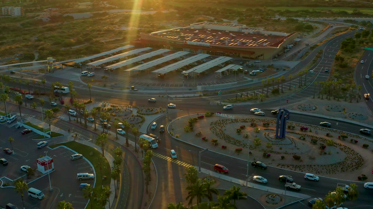 Elevated view of the San Jose Del Cabo roundabout with cars on the curved lanes, planted medians, palm trees, a large building in the background, and no visible people.