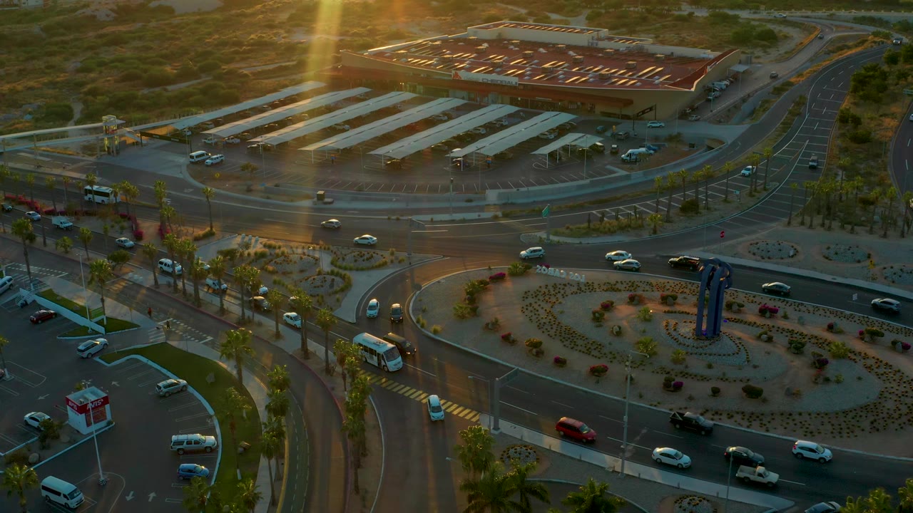 Aerial frame of the San Jose Del Cabo circular intersection with moving vehicles, palm-lined sidewalks, streetlights, landscaped islands, and no visible people.