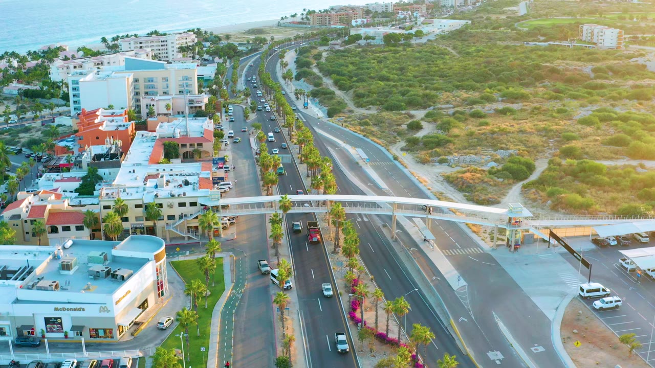 Aerial view over San Jose Del Cabo with cars on a multi-lane coastal road, a white pedestrian overpass, low buildings, green hillside, ocean in the distance, and no visible people.