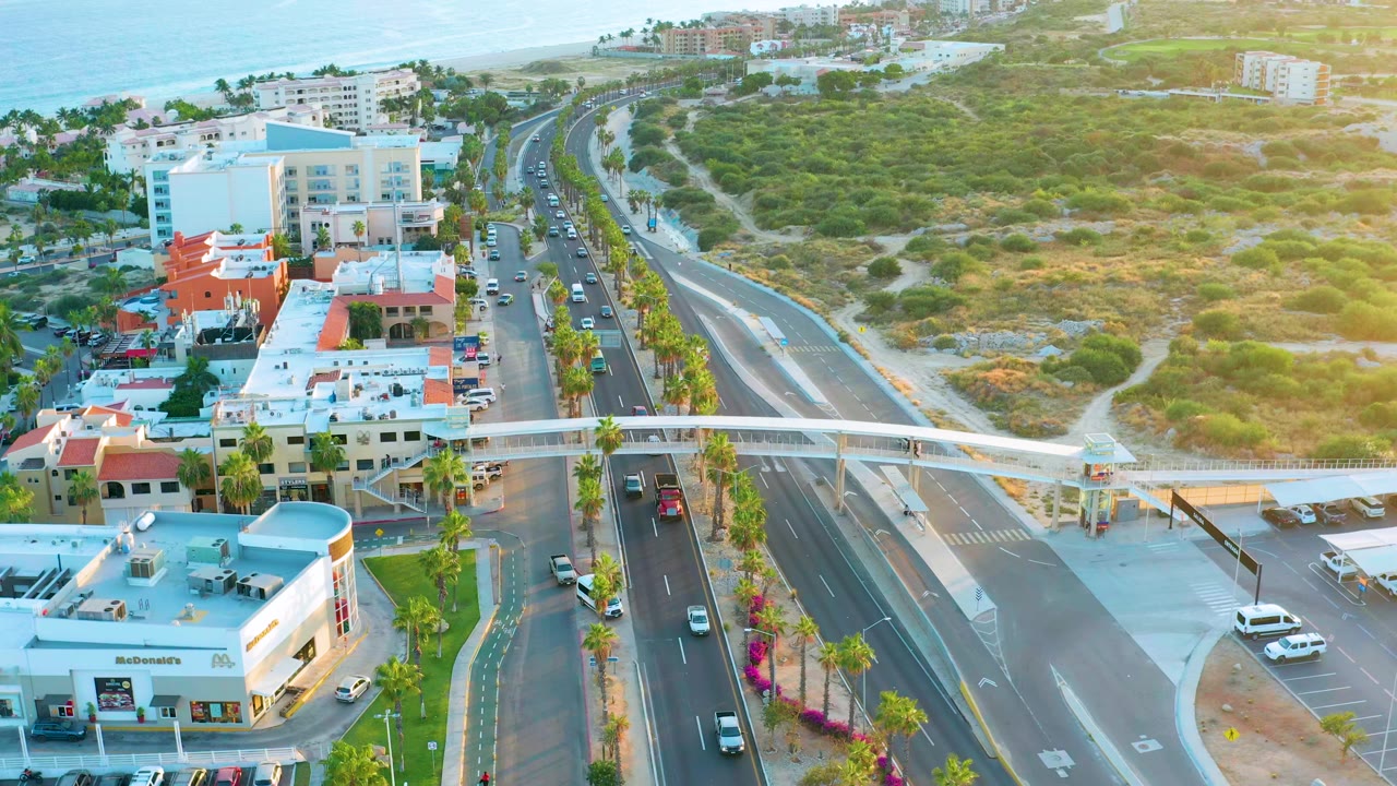 Aerial frame in San Jose Del Cabo showing traffic passing beneath and near a white pedestrian overpass, with palm trees, low commercial buildings, open hillside, ocean, and no visible people.