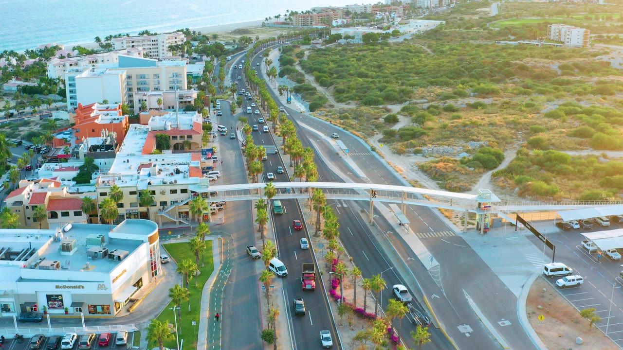 Aerial frame over San Jose Del Cabo shows cars moving along a divided coastal highway beside low buildings, palm trees, parking lots, hillside brush, beach, ocean, and no visible people.