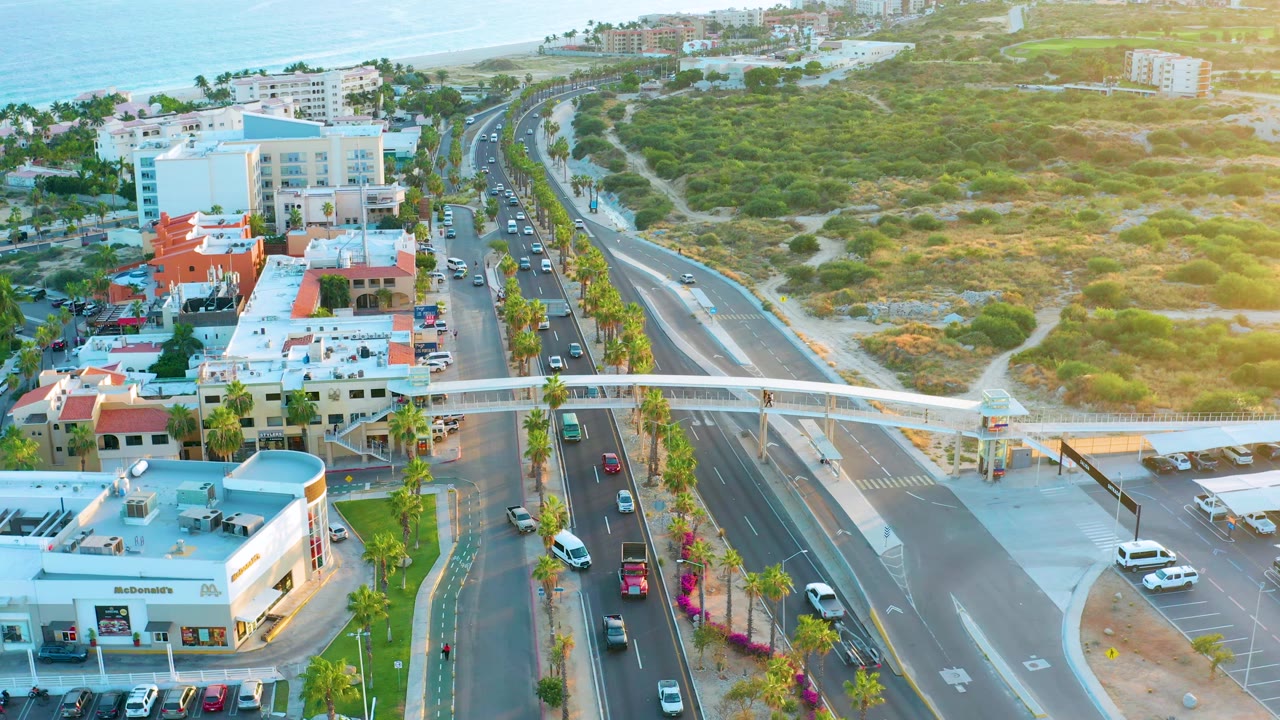 San Jose Del Cabo aerial view shows vehicles advancing beneath a white pedestrian bridge, with commercial buildings, parking areas, palm-lined lanes, hillside greenery, ocean, and no visible people.
