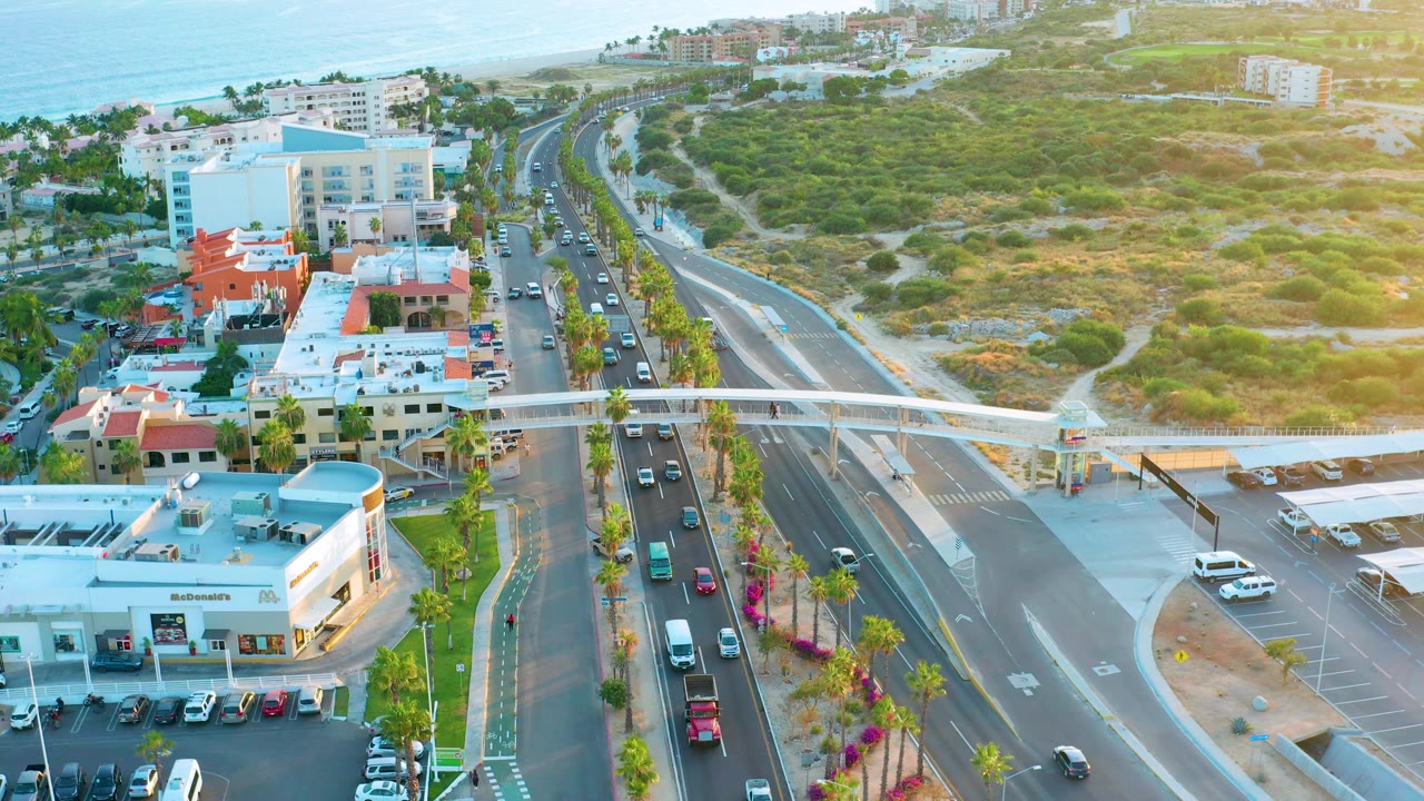 Aerial San Jose Del Cabo road view with traffic in both directions, commercial buildings in the lower frame, a white overpass across the road, hillside vegetation, ocean, and no visible people.