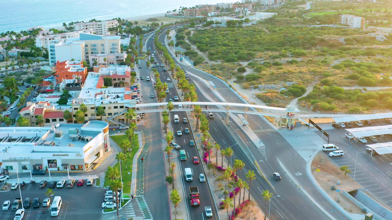 Aerial highway scene with palm-lined road edges, cars, buildings, and green hills.