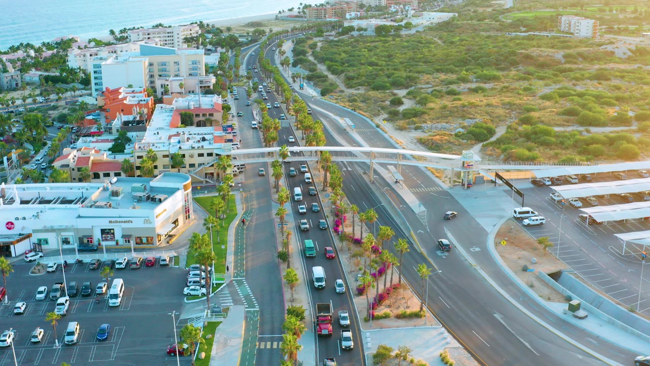 Opening aerial frame over San Jose Del Cabo showing slow traffic on a multi-lane road, a parking lot, low buildings, a pedestrian overpass, coastal scrubland, ocean in the distance, and no visible people.