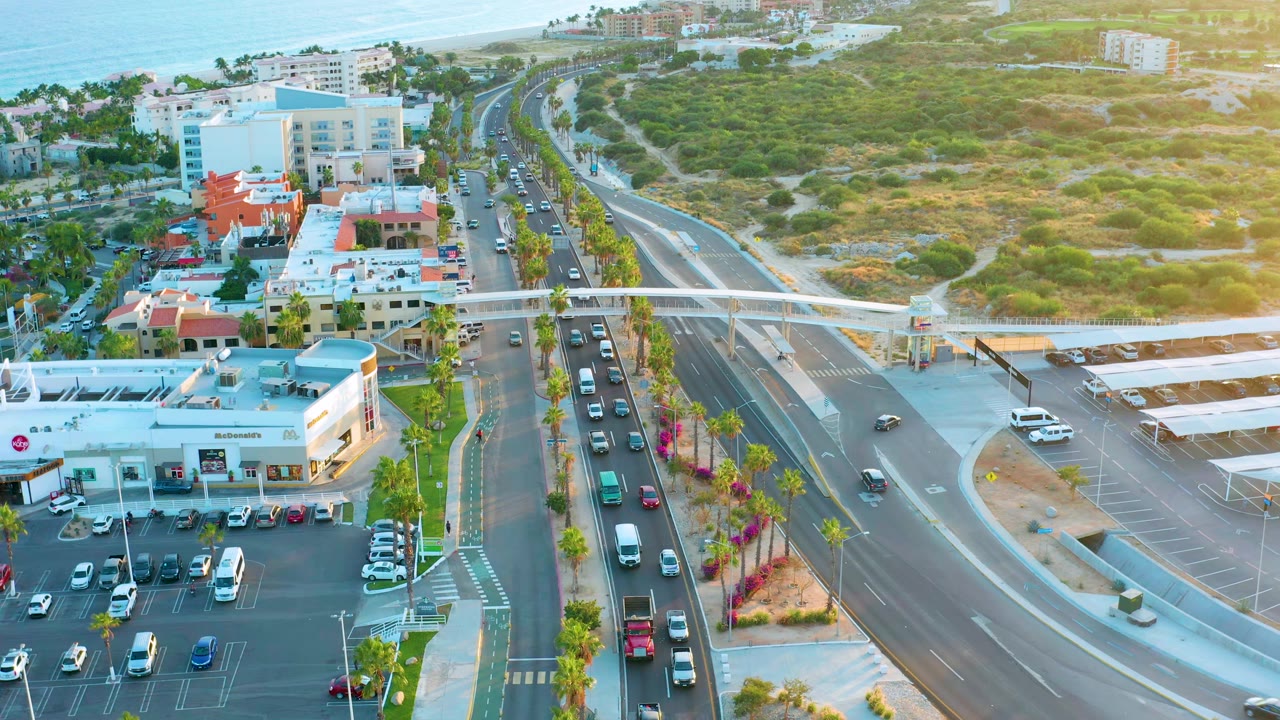 Drone view of cars on a divided road beside parking lots and coastal buildings.