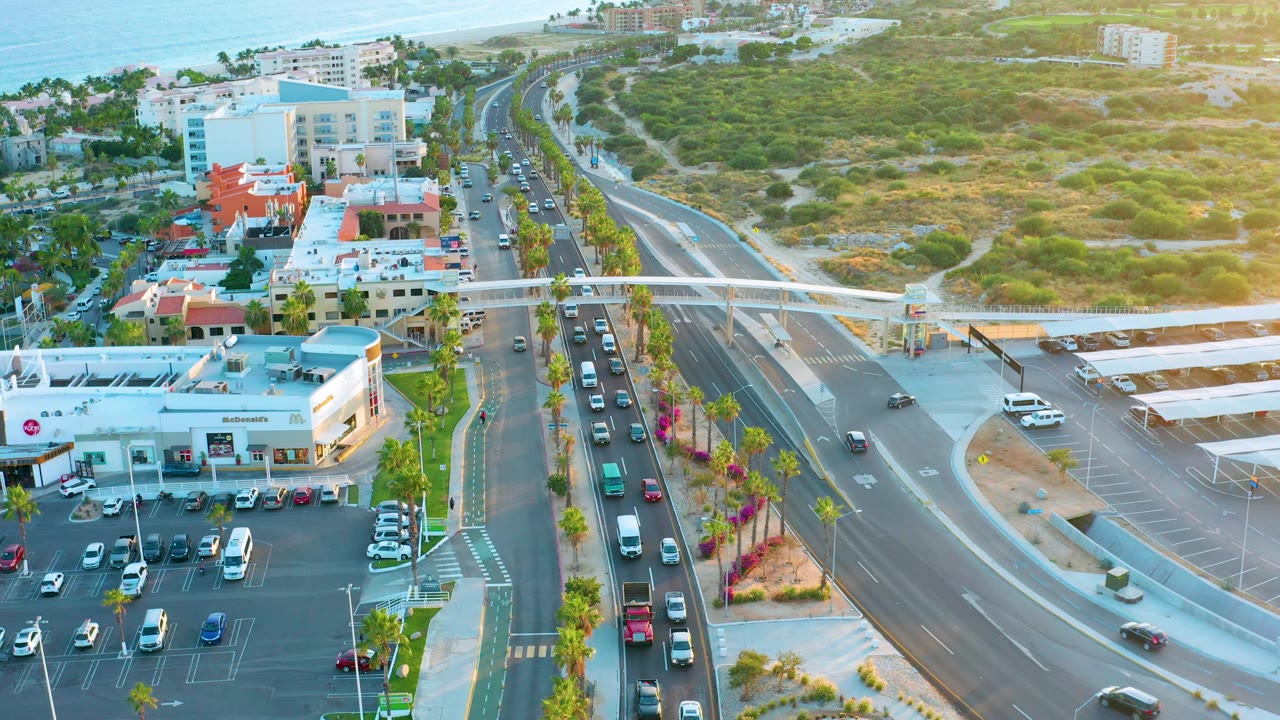 San Jose Del Cabo aerial frame showing a pedestrian overpass across the road, vehicles moving below, a curved side road, parking lots beside commercial buildings, green coastal scrub, ocean background, and no visible people.