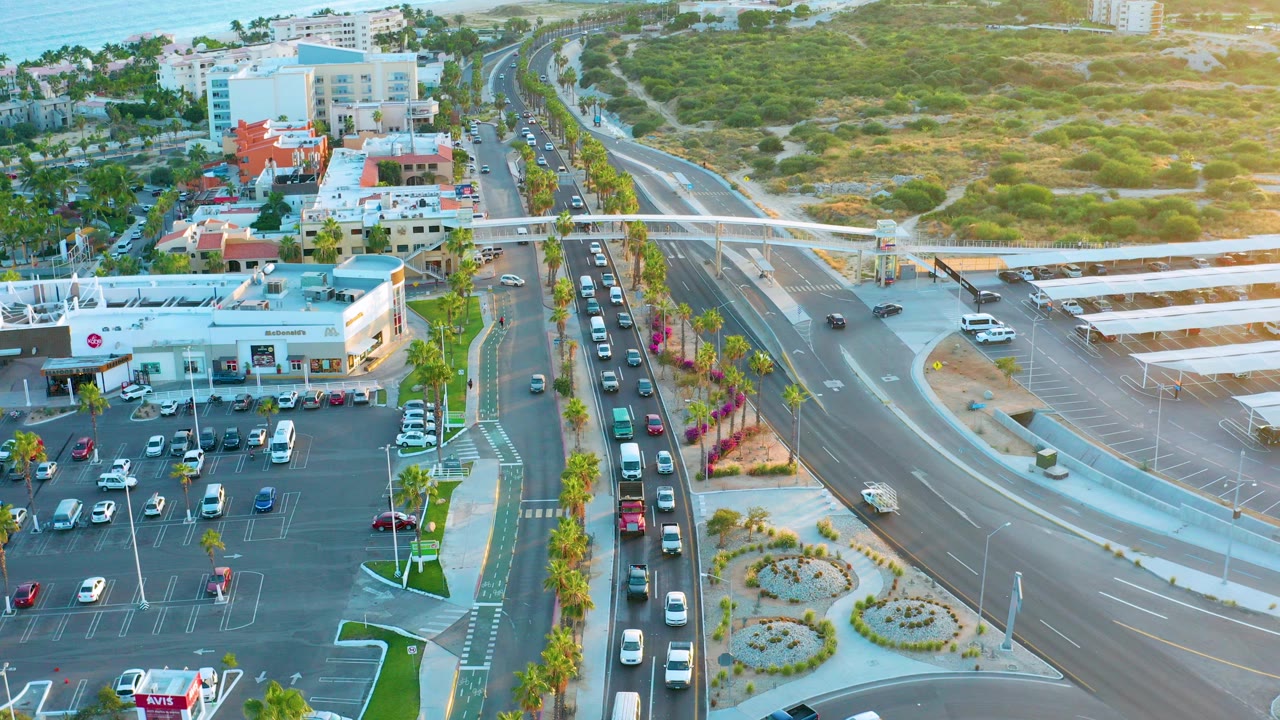 Aerial frame over San Jose del Cabo showing a multi-lane road intersection, a commercial parking lot, palm trees, low buildings, and no visible people.