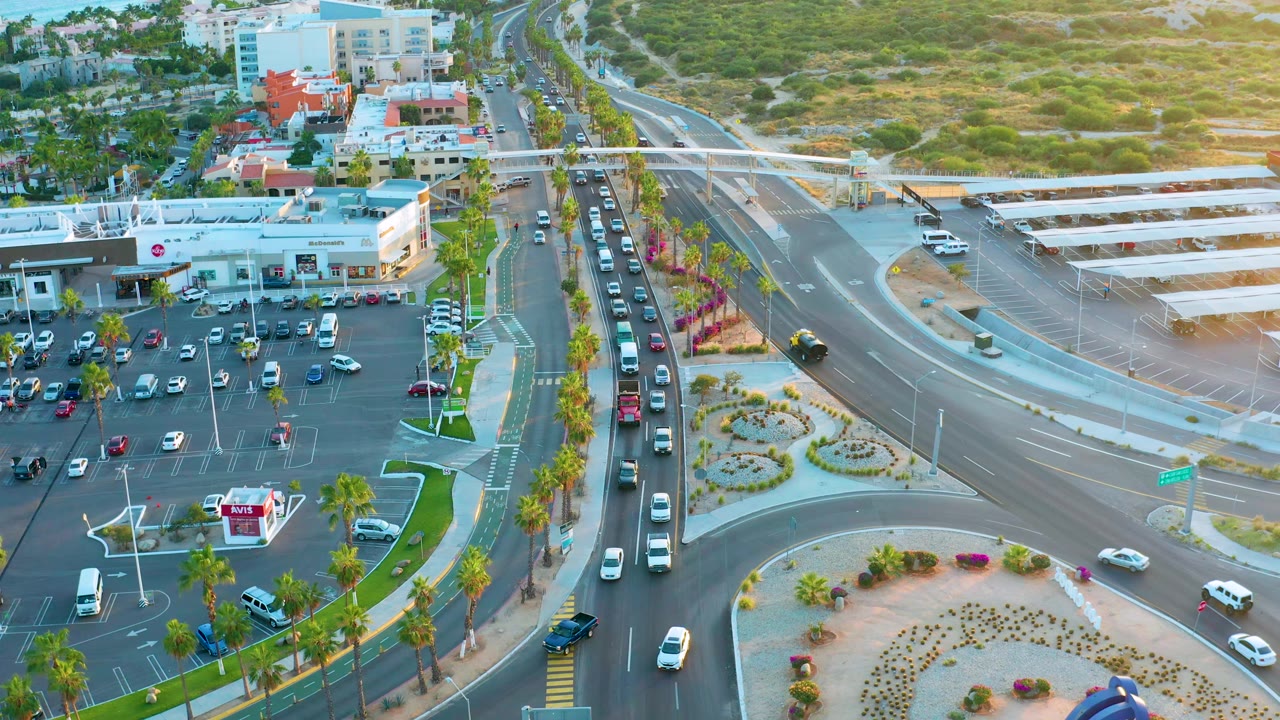 Second aerial frame over San Jose Del Cabo with cars shifting through the intersection, rows of parked vehicles at the commercial lot, palm landscaping, and no visible people.