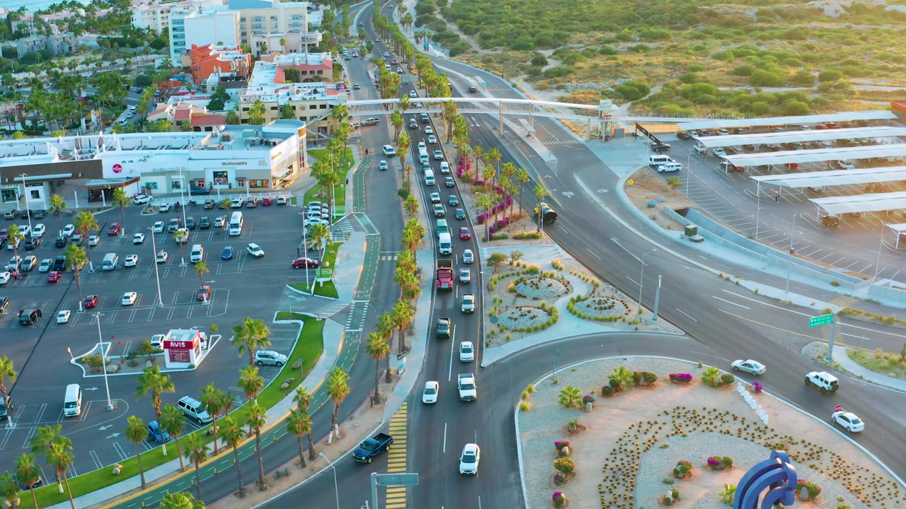 Aerial San Jose Del Cabo traffic frame with vehicles spread across curved lanes, a busy parking lot to the left, palm rows along the road, and no visible people.