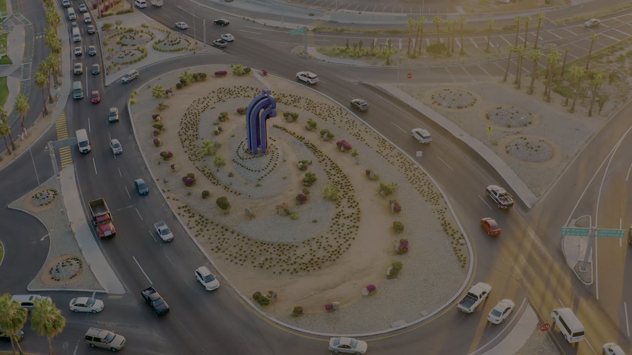 Aerial frame of a San Jose del Cabo roundabout with cars and vans moving around a planted central island with a blue sculpture, no visible people.