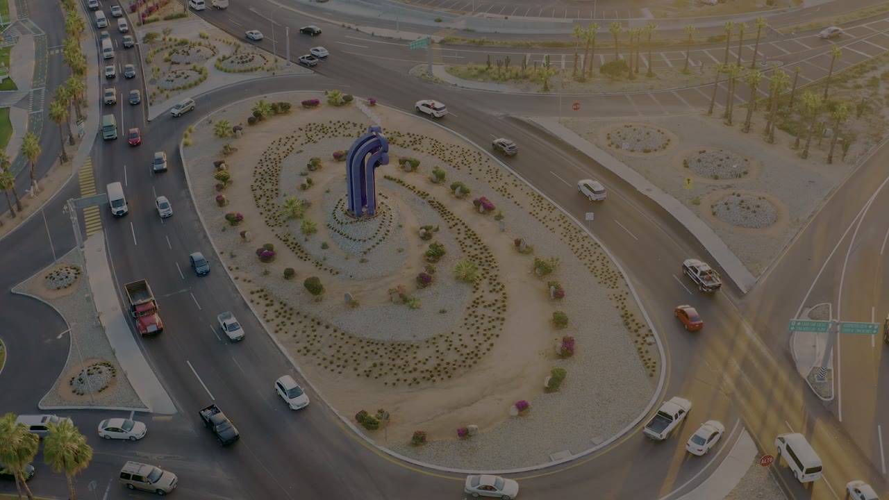 High aerial view in San Jose del Cabo showing cars and a white van circulating beside a desert-planted traffic circle with the blue sculpture near center, no visible people.