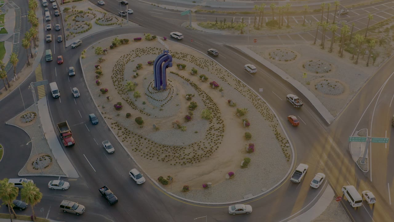 Aerial San Jose del Cabo traffic frame with the roundabout island shifted high in view, several cars entering and exiting, and the blue central sculpture visible, no visible people.