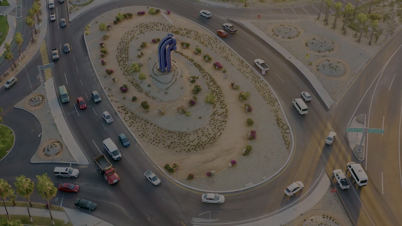 Aerial San Jose Del Cabo traffic frame with a white van near the right feeder road, cars on the left lanes, the planted roundabout island, and no visible people.
