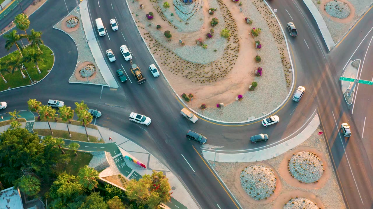 Overhead aerial frame in San Jose Del Cabo showing cars entering and exiting a landscaped roundabout with desert plantings, palm trees, and no visible people.