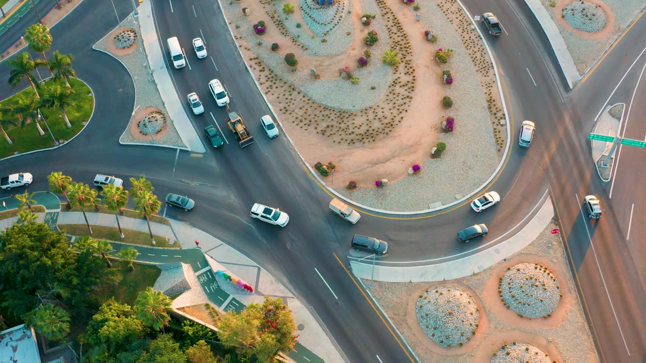 San Jose Del Cabo overhead frame showing cars spread around the roundabout, pale sidewalks, decorative desert beds, palm trees, and no visible people.