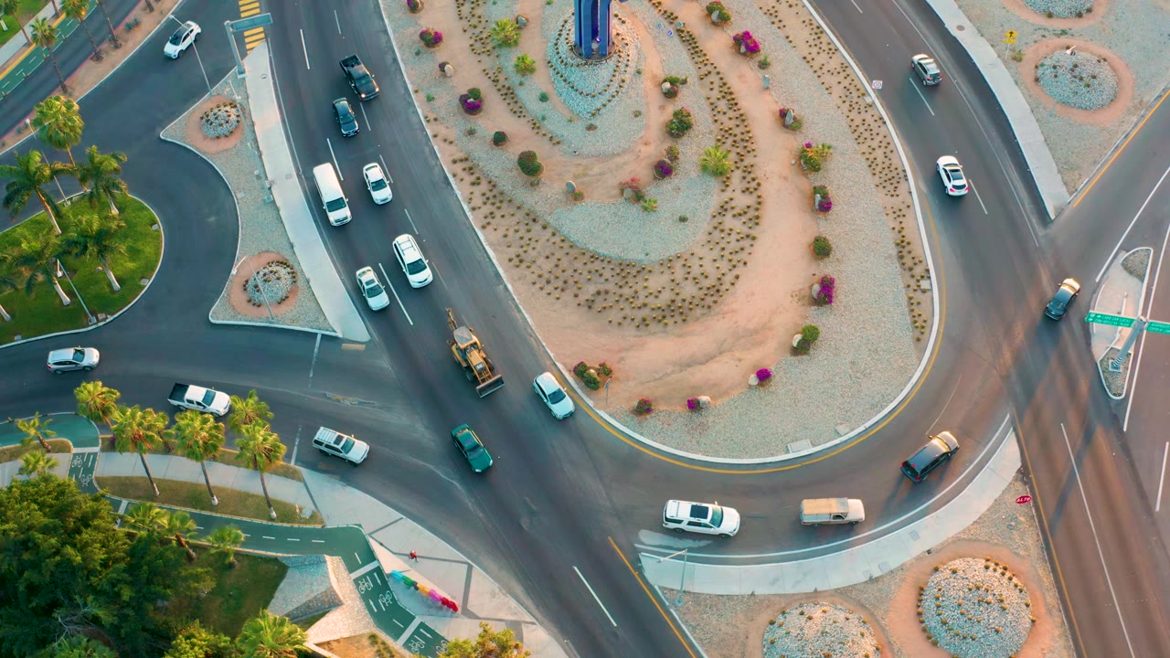 Overhead frame from San Jose Del Cabo showing white cars, dark vehicles, and a small truck moving around the curved lanes beside a planted central island, with no visible people.