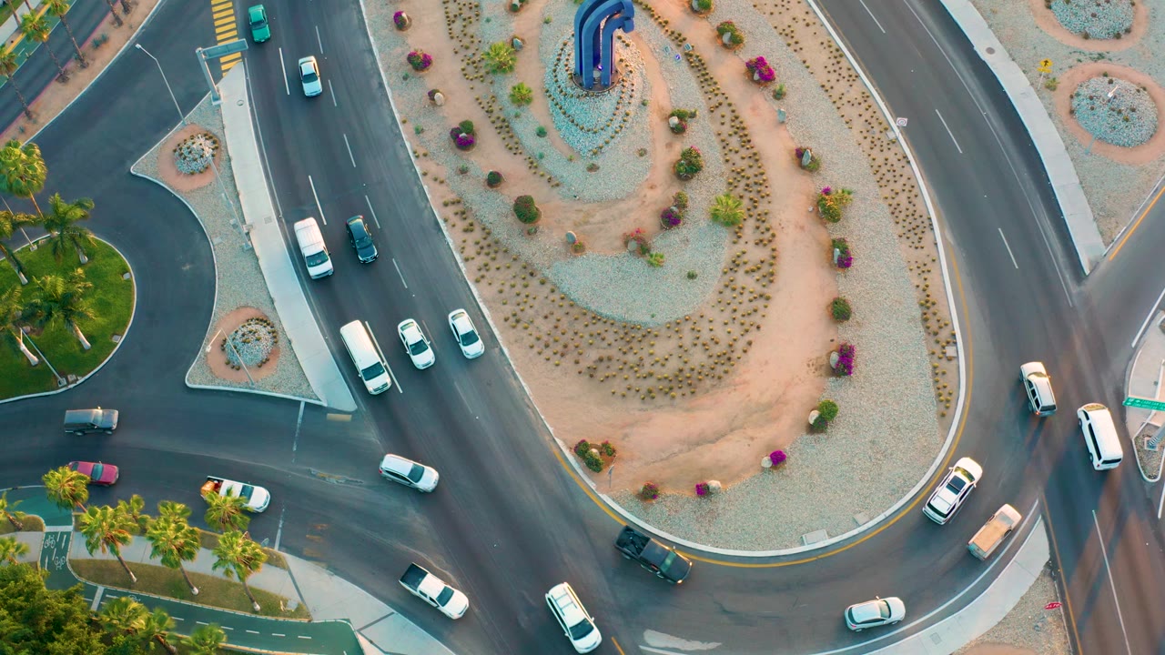 Top-down frame of the San Jose Del Cabo roundabout with cars spaced across the left and lower lanes, a sweeping central island of gravel and plants, and no visible people.