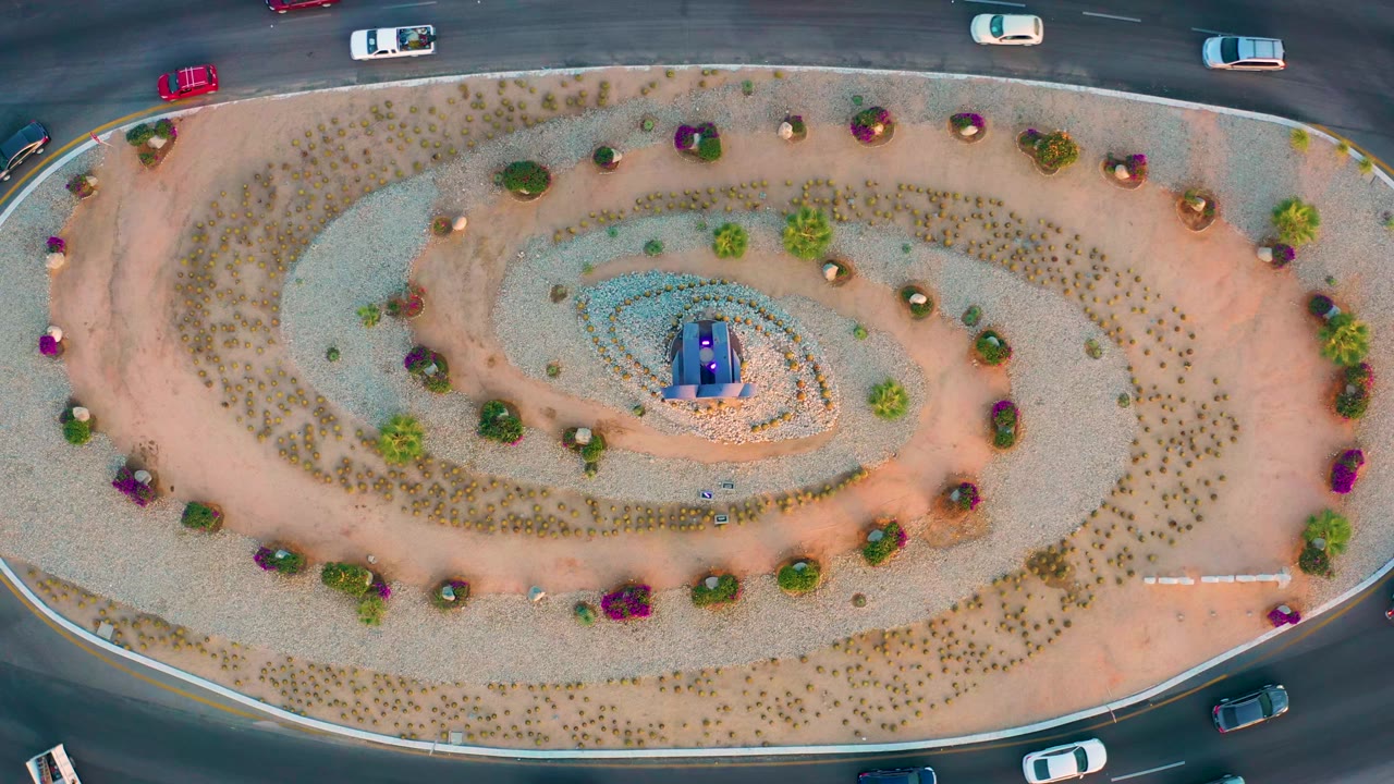 Aerial frame from San Jose Del Cabo with the oval spiral garden centered and several vehicles moving along the dark roads at the top and bottom edges, with no visible people.