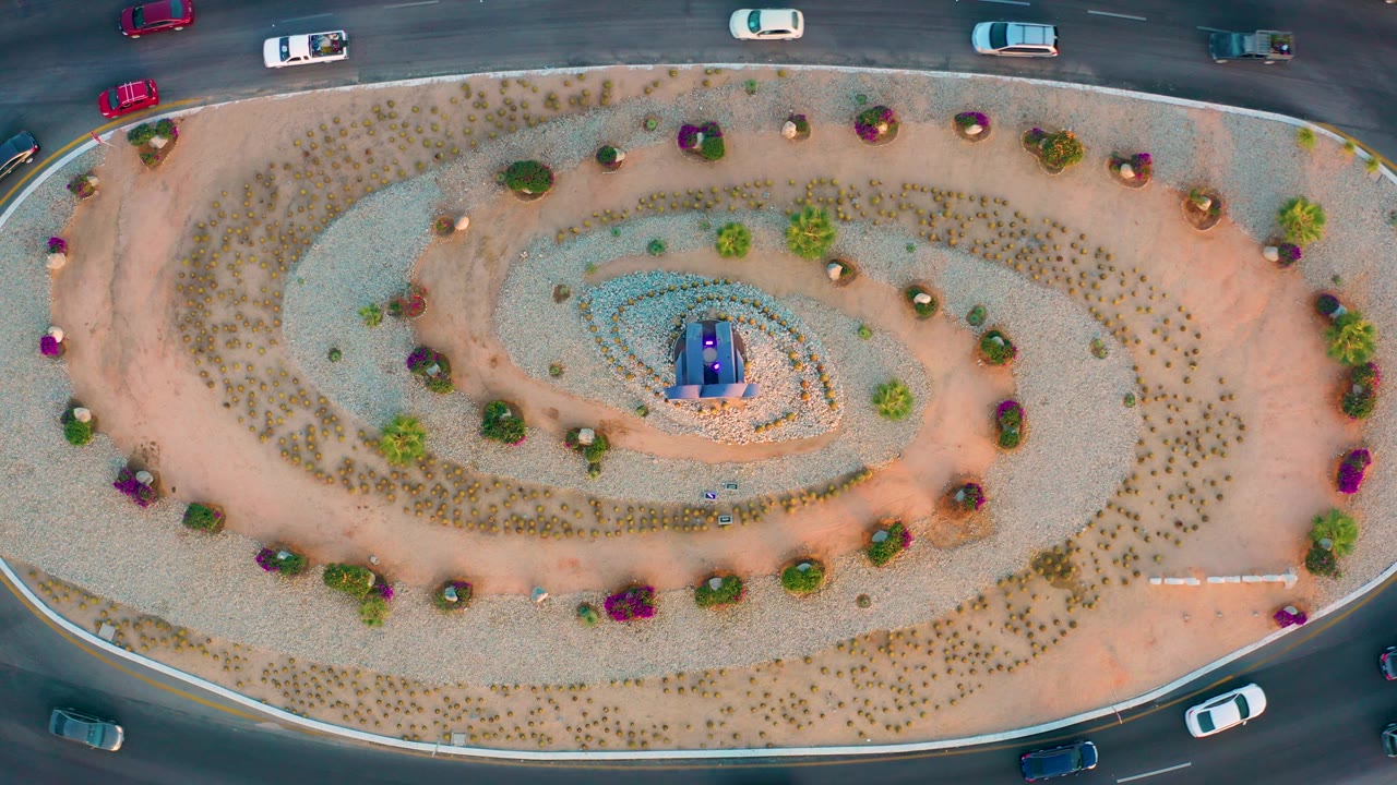 San Jose Del Cabo overhead frame with vehicles visible on the upper and lower roads around the oval landscaped roundabout, plus the blue and purple center detail and no visible people.