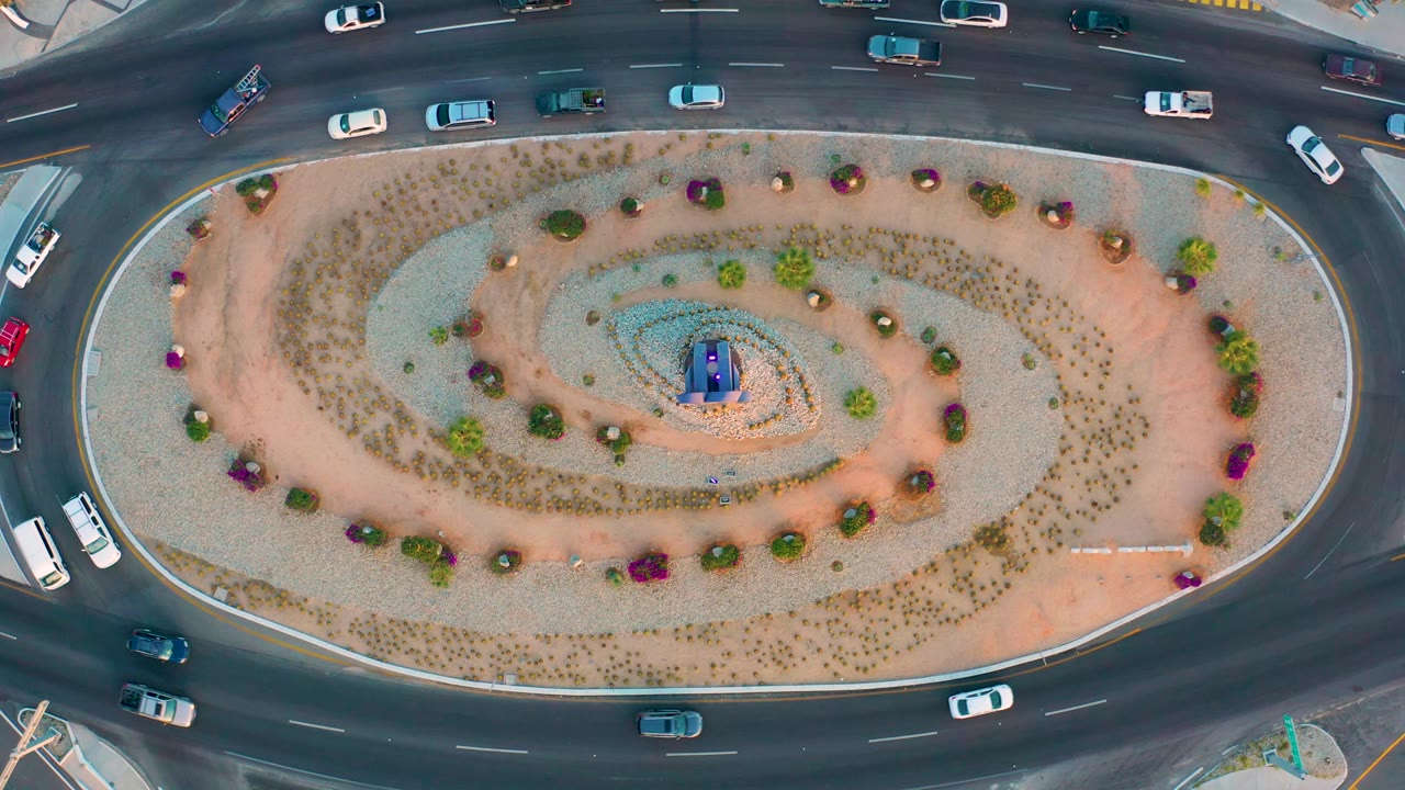 Top-down San Jose Del Cabo view of a central oval garden with spiral gravel beds, planted shrubs, and cars spaced along the surrounding lanes, with no visible people.