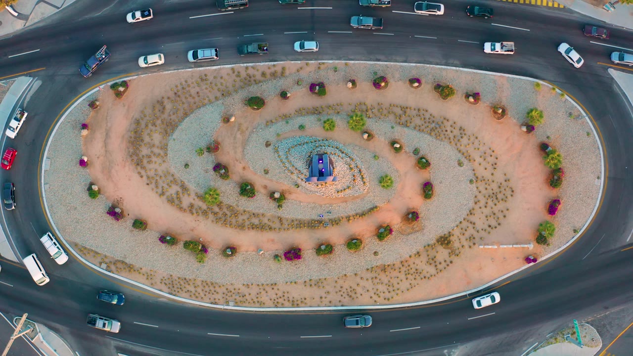 Aerial frame over San Jose Del Cabo with cars, small vans, and a bus-like vehicle moving around a landscaped oval median and no visible people.