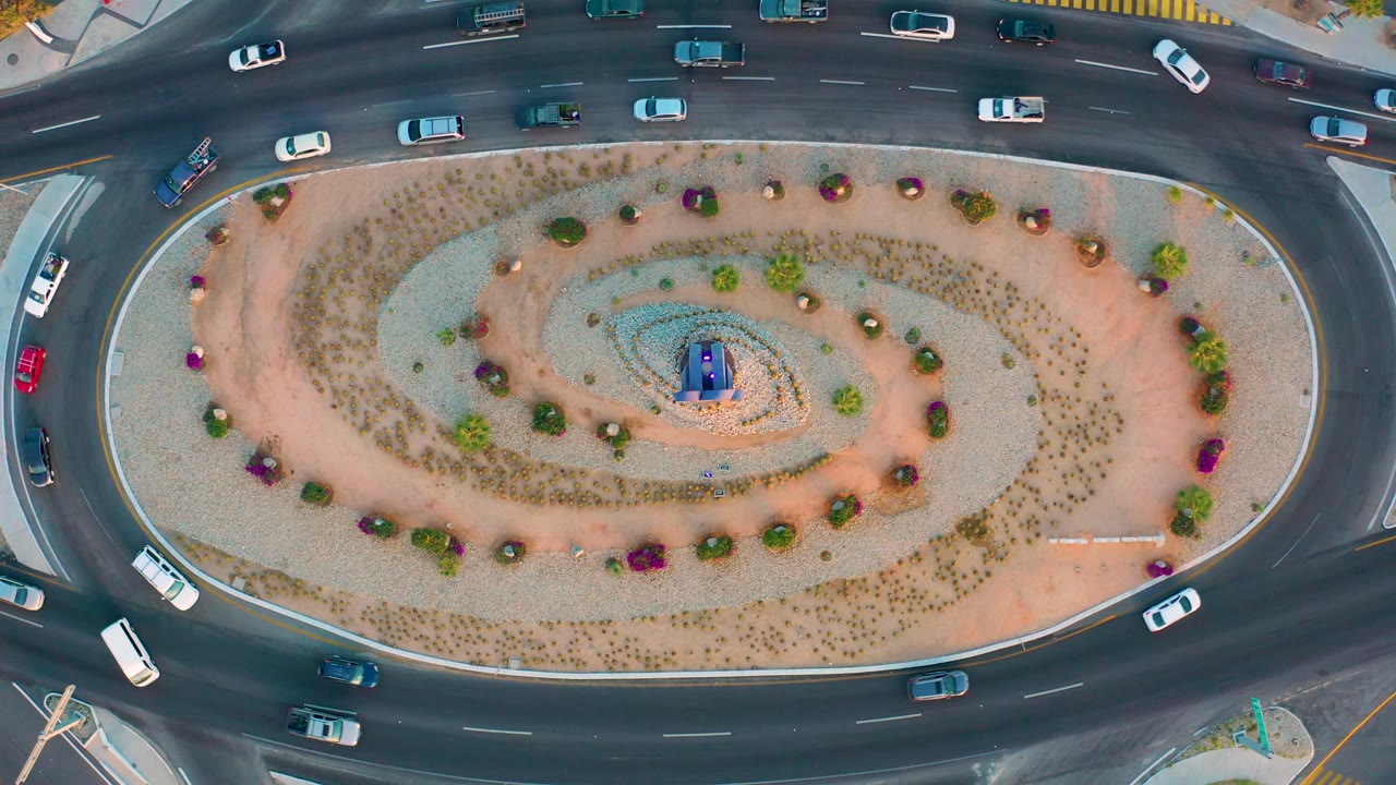 Overhead San Jose Del Cabo frame showing vehicles spread around the dark asphalt ring, an oval planted center, and a pale sidewalk edge, with no visible people.