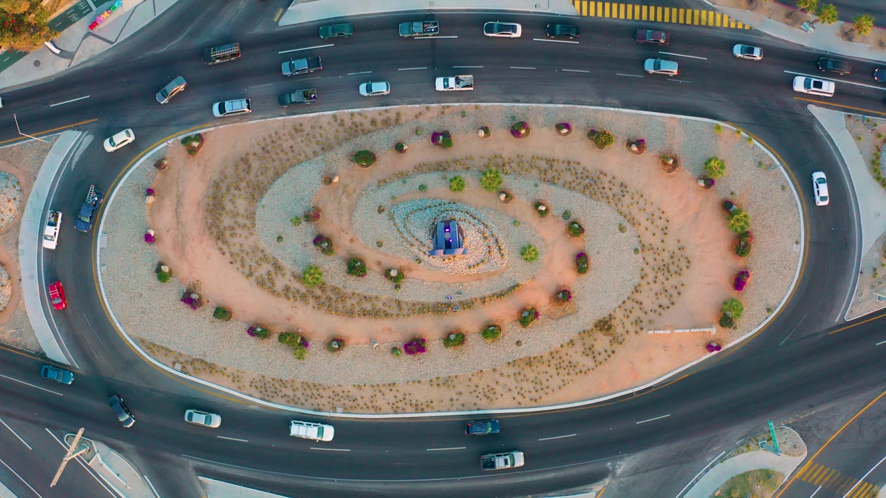 Top-down aerial frame of a multi-lane roundabout in San Jose Del Cabo with cars on curved lanes, a planted oval center island, sidewalks at the edges, and no visible people.