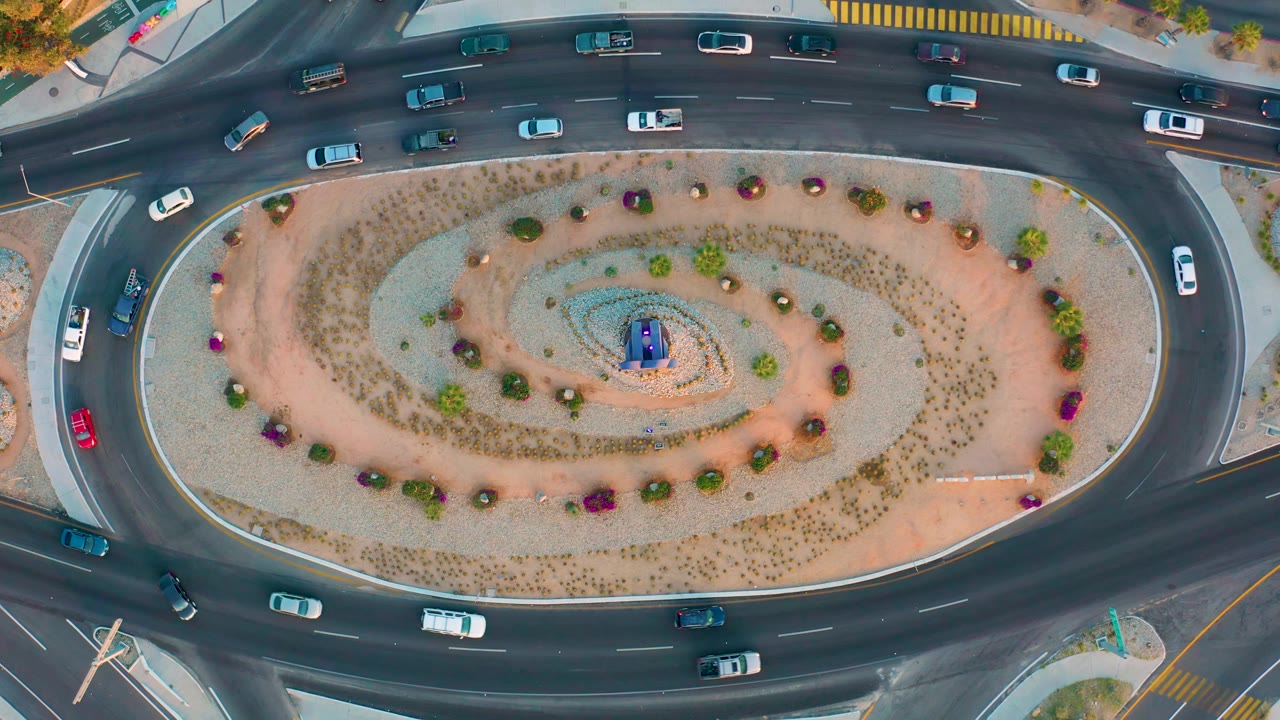 Overhead aerial frame in San Jose Del Cabo showing more vehicles spaced along the upper curve of the roundabout, with the oval gravel island and planted spiral pattern centered below, and no visible people.
