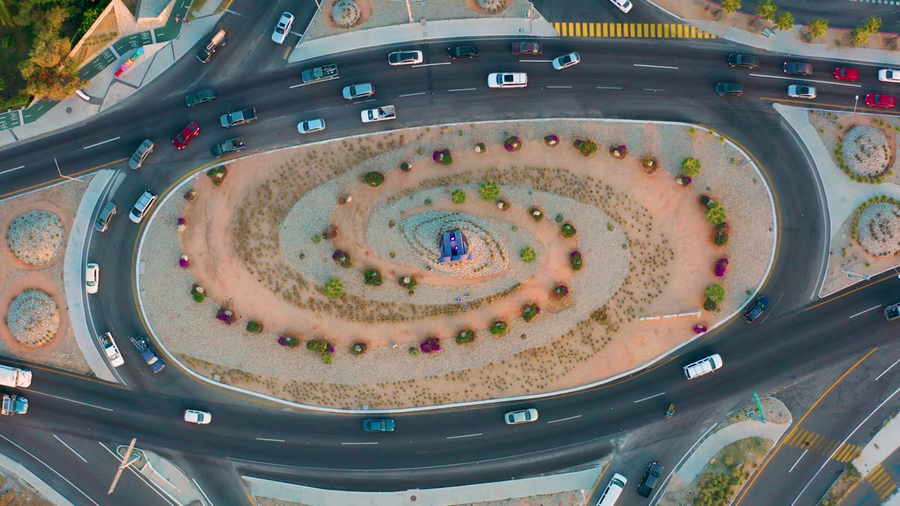 Overhead frame of a landscaped roundabout in San Jose del Cabo with cars moving through multiple lanes, sidewalks and crosswalks at the edges, and no visible people.