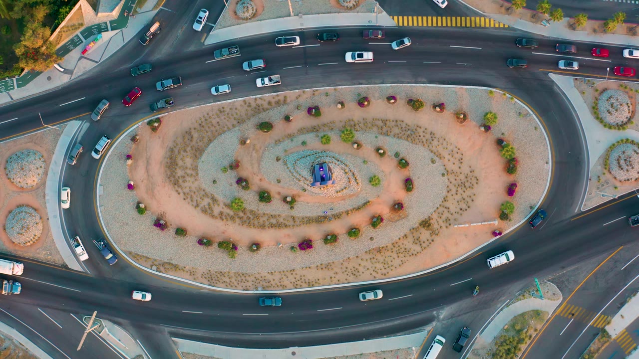 Aerial view in San Jose del Cabo showing several cars and larger vehicles spaced around an oval planted traffic island, with no visible people in the road scene.