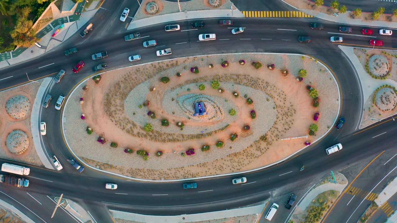 Top-down San Jose del Cabo frame with cars positioned on entry and exit lanes around a landscaped center, plus sidewalks, curbs, and no visible people.