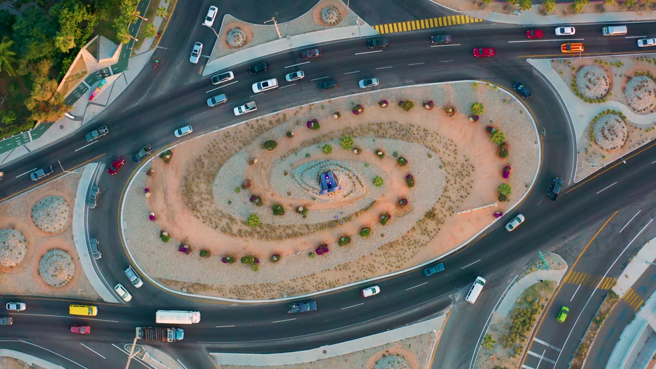 Aerial frame over San Jose Del Cabo showing cars distributed around the curved lanes, pale landscaping inside the roundabout, and no visible people.