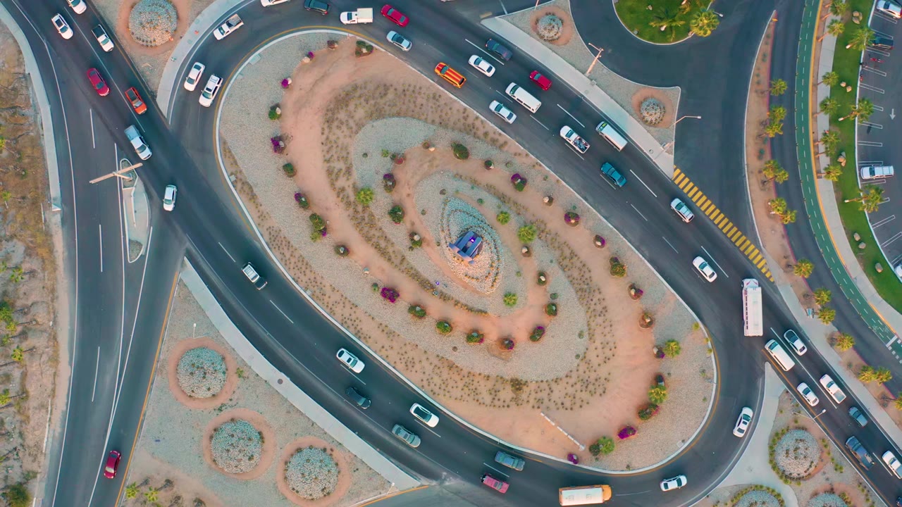 Overhead frame in San Jose del Cabo showing more vehicles spaced around the roundabout, including a long white bus near the right side and no visible people.
