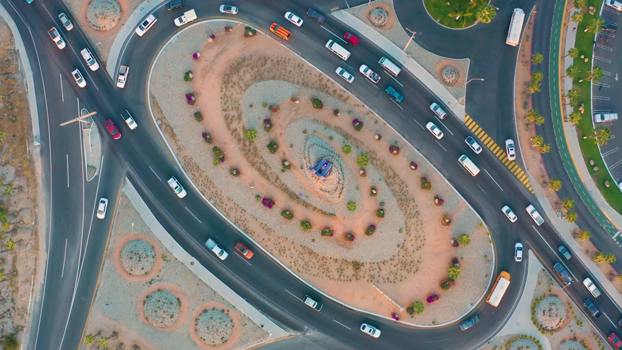 Top-down San Jose Del Cabo traffic view with white, red, blue, and green vehicles circling the roundabout beside planted medians and no visible people.