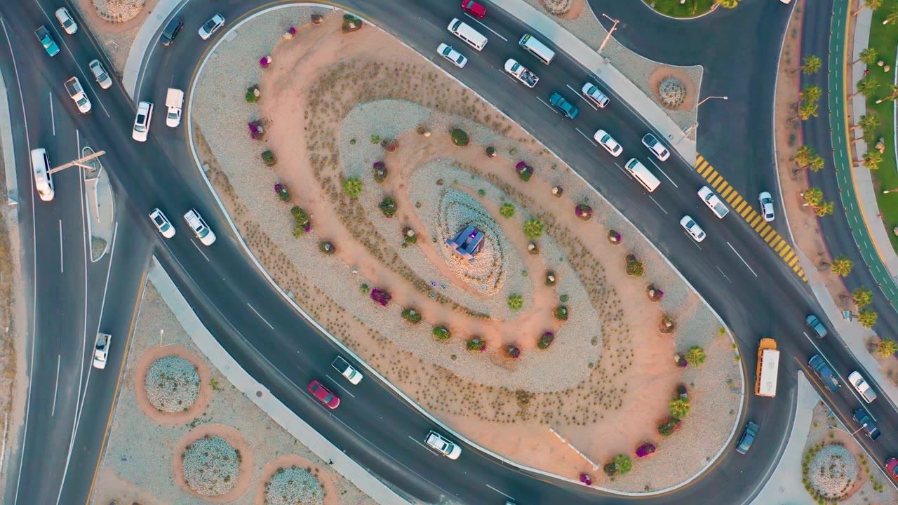 Aerial frame in San Jose Del Cabo showing white and blue cars spaced around the curved lanes of the roundabout, with no visible people.