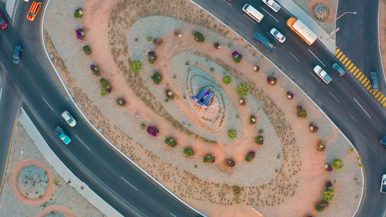 Aerial top-down frame in San Jose Del Cabo showing multiple cars and a bus around the roundabout, with yellow crosswalk markings near the upper road and no visible people.