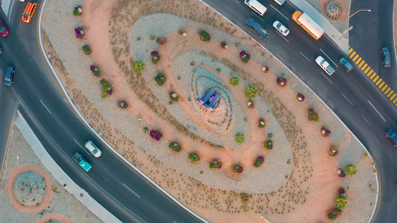 Top-down San Jose Del Cabo traffic circle with cars spaced across the upper and left roads, a planted gravel island, blue structure, and no visible people.