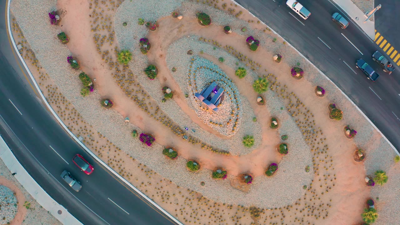 Aerial frame in San Jose Del Cabo showing vehicles spaced along the road beside concentric gravel and planting rings, with the central garden feature near the middle and no visible people.