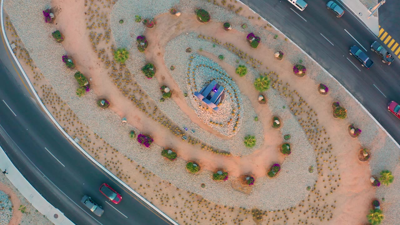 Straight-down view of the San Jose Del Cabo roundabout with several cars along the upper and left road lanes, landscaped gravel circles, bright flowering shrubs, and no visible people.