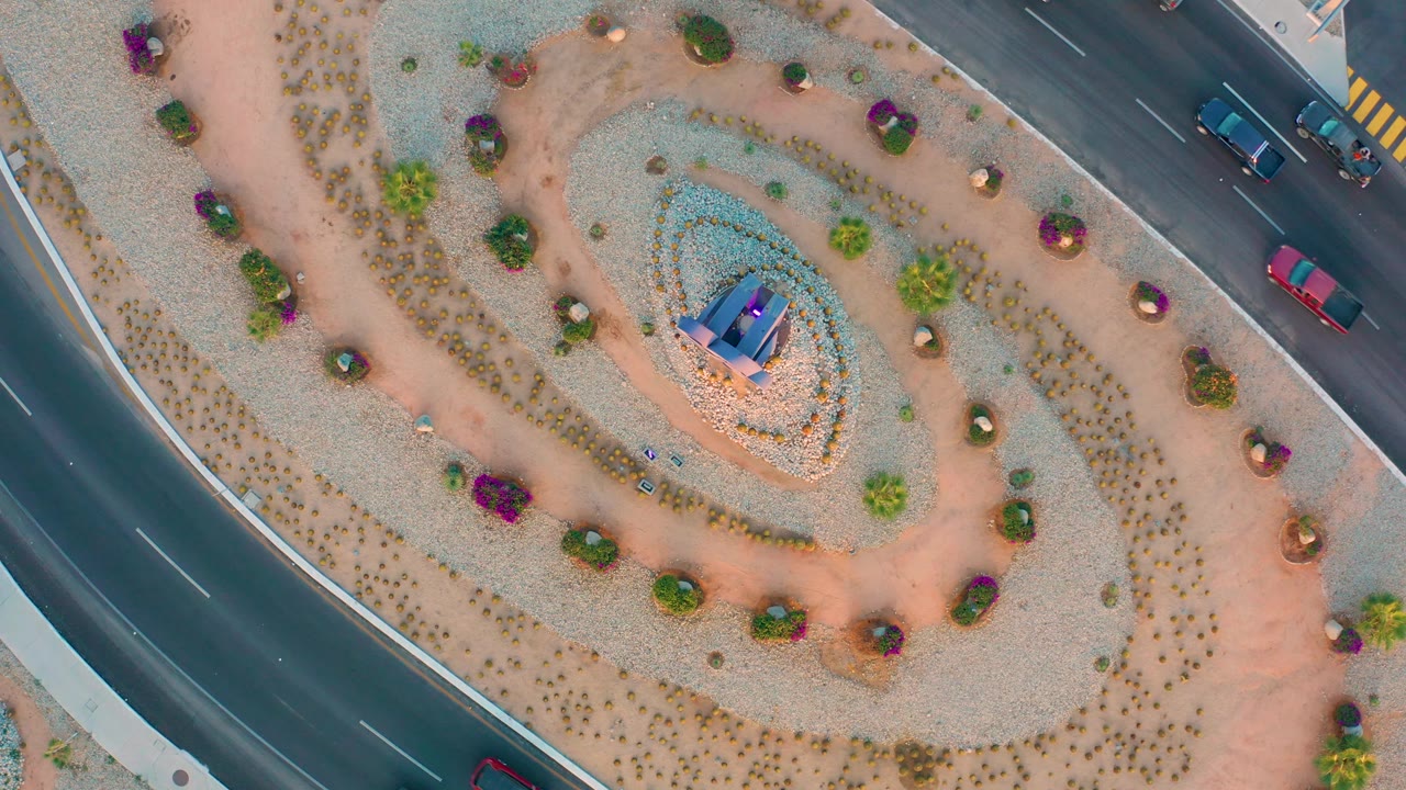 Top-down frame from San Jose Del Cabo with the central garden feature low in the island, curved tan paths around pale gravel, cars on both road edges, and no visible people.