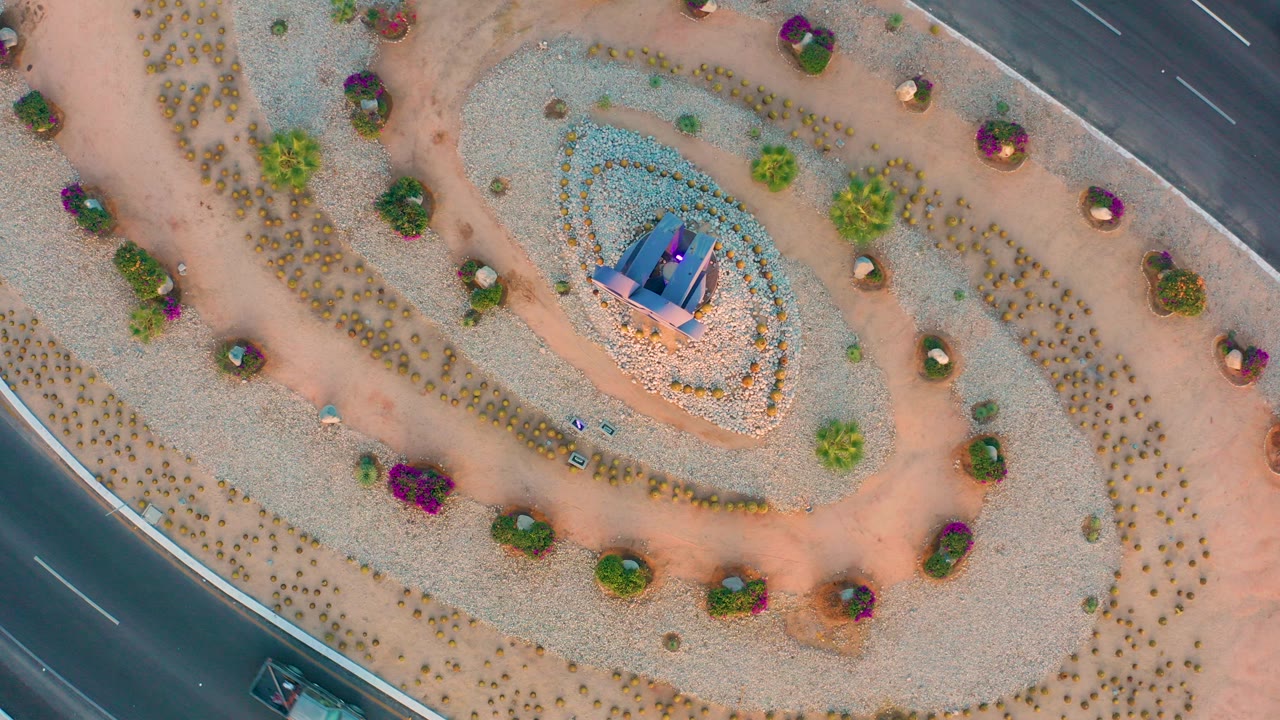 Top-down aerial frame in San Jose Del Cabo showing a spiral gravel garden around a central monument, sandy paths, small shrubs, purple flowers, road edges, and no visible people.