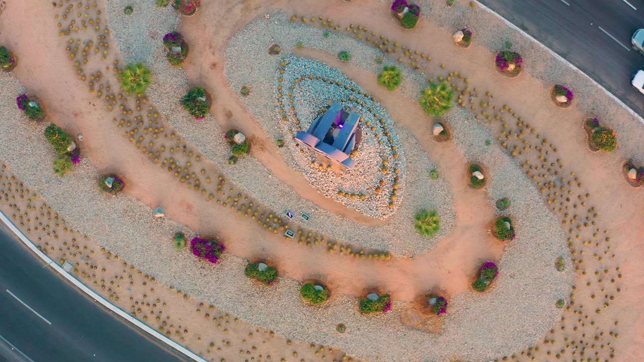 Aerial frame over San Jose Del Cabo with a triangular road corner at lower left, a spiral garden bed around the central monument, scattered shrubs, purple flowering plants, and no visible people.