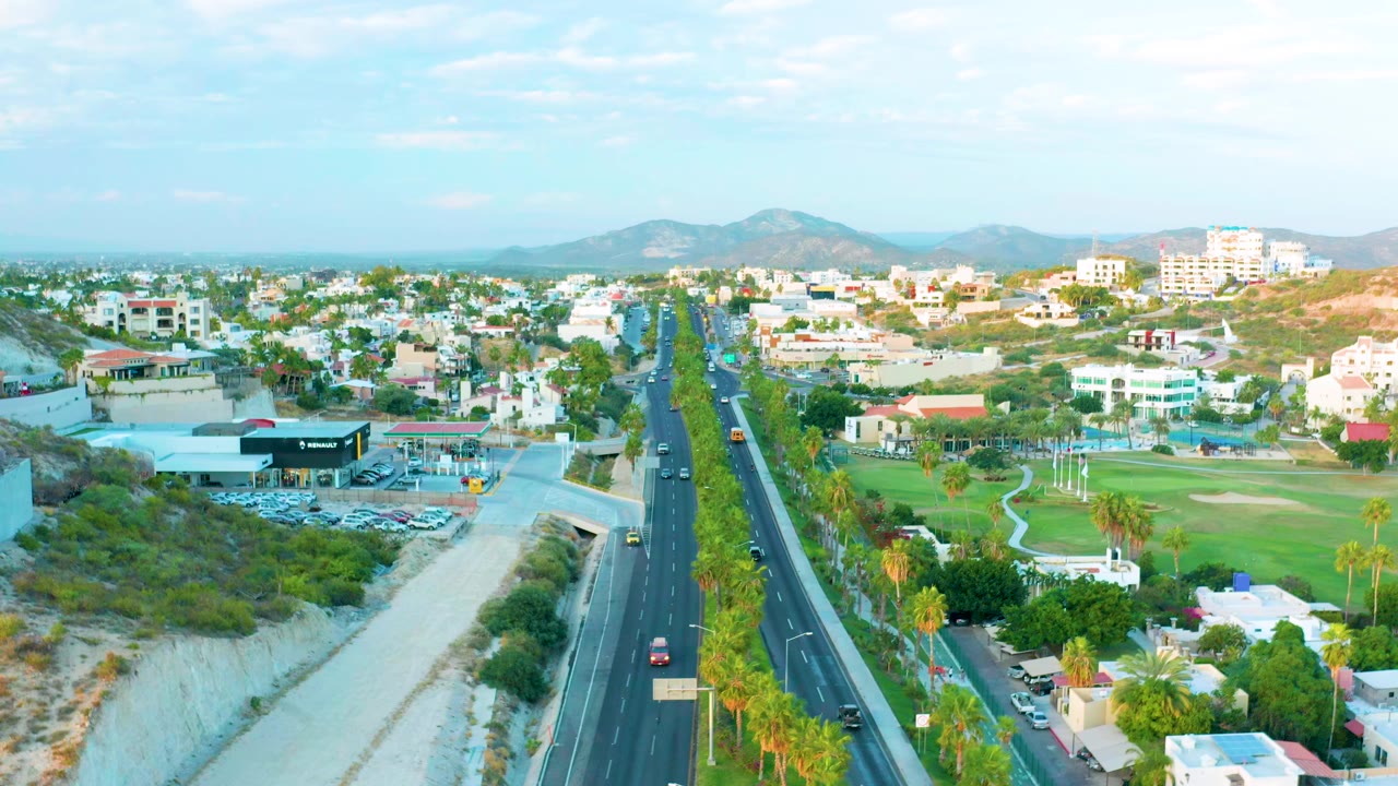 Aerial frame from San Jose Del Cabo showing a multi-lane road through the center, dry riverbed at left, green field with palms at right, distant buildings and mountains, and no visible people.