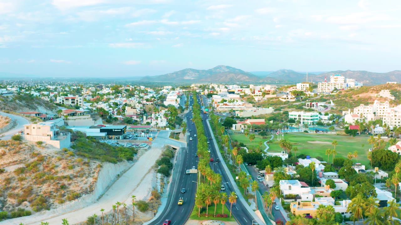 One second into the clip, the San Jose Del Cabo aerial holds the road in the center with cars spaced along the lanes, palms and green grounds below, dry slopes beside it, and no visible people.