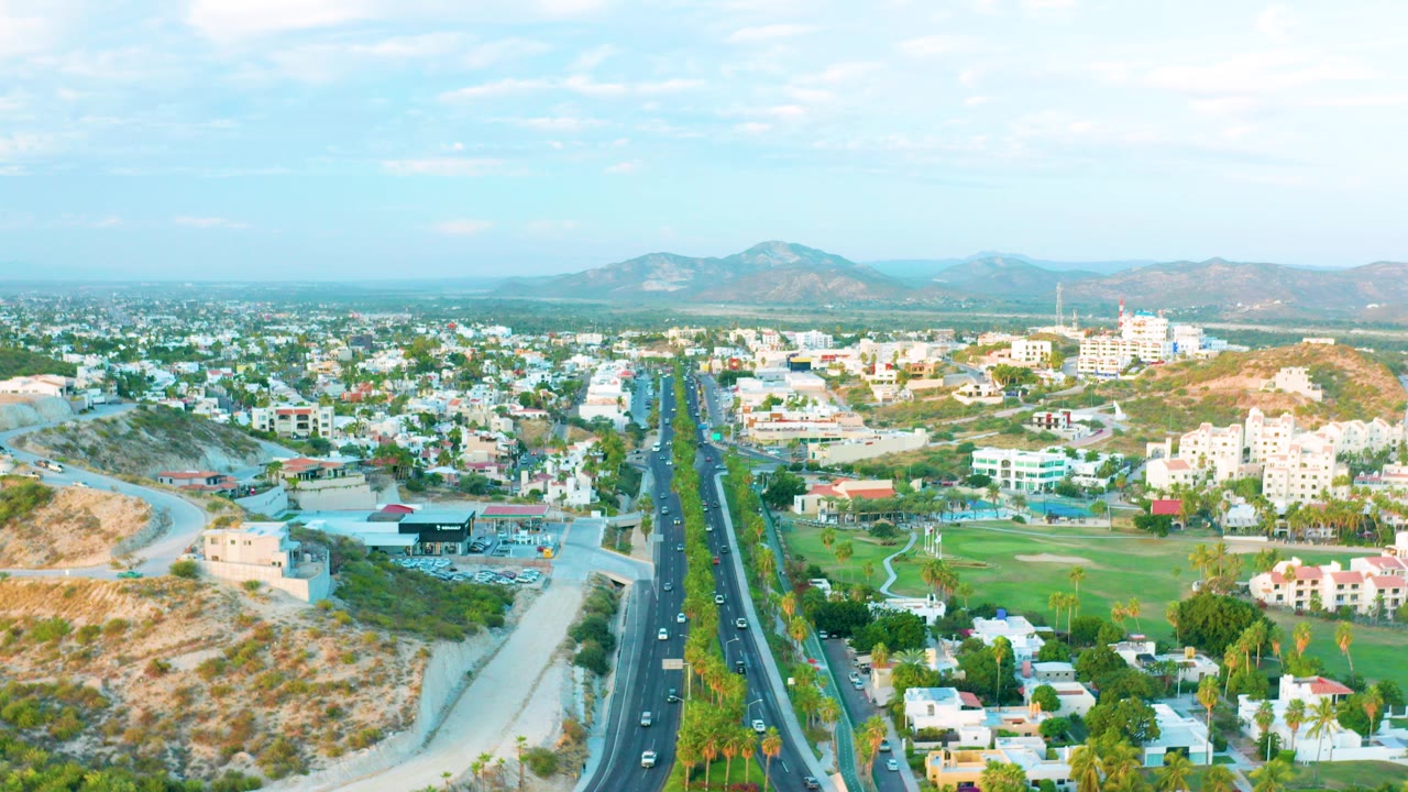 Opening aerial frame over San Jose Del Cabo showing a divided road with cars, low buildings, green grounds, dry hills, distant mountains, and no visible people.